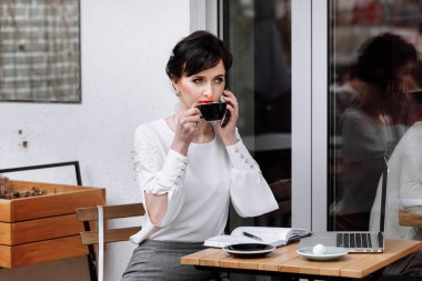 İş kadını akıllı telefonuyla konuşuyor, dizüstü bilgisayarla çalışıyor ve şehir merkezindeki terasta kahve içiyor. Müşterilerle uzaktan iletişim. Kahve molası, iş konsepti. Açık havada çalışmak.