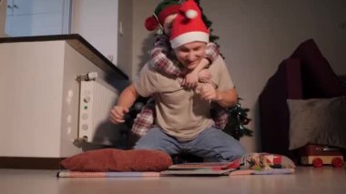 Neşeli çocuk kız Noel Baba şapkalı mutlu babayla Noel ağacının yanında oynaşırken eğleniyor. Kış tatilini evde kutlayan samimi ve aktif bir aile. Aileler birlikte eğlenir..