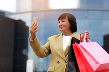 Alışveriş çantaları ve akıllı telefonları olan şık yetişkin kadın blogcu alışveriş merkezi yakınlarındaki sosyal ağlarda takipçilerle iletişim kuruyor. Tüketim, satış, zengin hayat, sanal hayat kavramı. Kara Cuma.