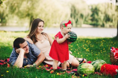 Aile piknikte. Anne, baba ve küçük kız parkta battaniyenin üzerinde oturuyorlar. Yaz tatili kavramı. Anneler günü, babalar günü, bebekler günü. Birlikte vakit geçirmek.