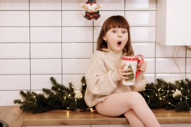 Şaşırmış küçük bir kız elinde sıcak bir içecekle bir fincan tutuyor ve Noel için Noel süslenmiş bir mutfakta yeni yılı bekliyor. Evde sıcak bir kış günü. Noel tatilinde mutlu bir aile.