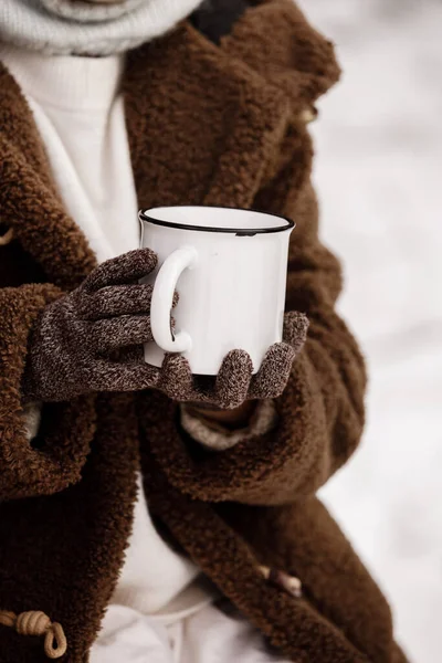 Cropped photo of little girl in warm clothes and mittens holding a big white metal cup of hot drink in hands in snowy forest. Adventure with family on picnic on winter holiday vacation.