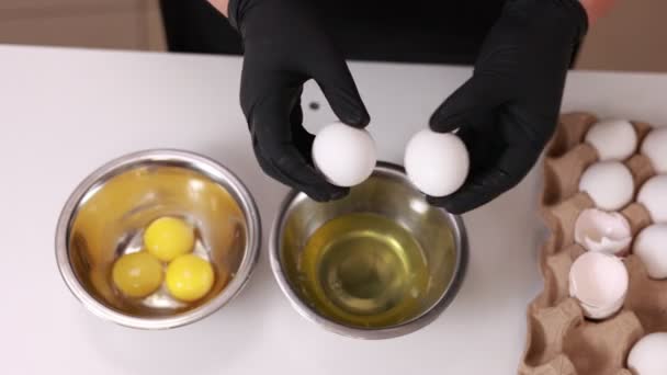白い背景に金属ステンレス混合ボウルの上に黒い手袋で手で白い生の殻の卵の黄身と白を分離 ベーカリーの材料や調理用の紙箱に卵 — ストック動画