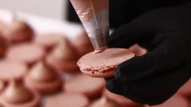 Making french desert macarons. Chef hands in black gloves with confectionery bag add cream to the ready halves of brown chocolate macaroons at pastry shop. cooking, food and baking concept