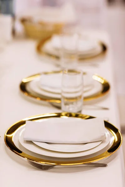 Banquet, restaurant. Elegant festive minimalist table setting with gold-white plates, glasses, cotton napkins and cutlery on table with white tablecloth. Vertical photo.