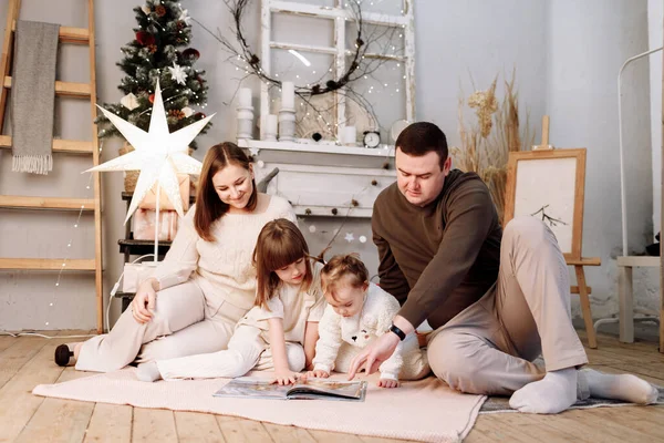 stock image Loving family. Mom, dad with little daughters read interesting book and having fun near decorated fir tree and fireplace with garlands. Merry Christmas and Happy New Year. Cozy winter evening at home.