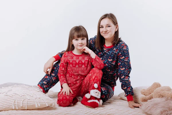 Happy family is hugging and having fun together. Smiling mom with little daughters in winter pyjamas in xmas prints are sitting on warm plaid on white background. Mothers day, baby day. New Year.