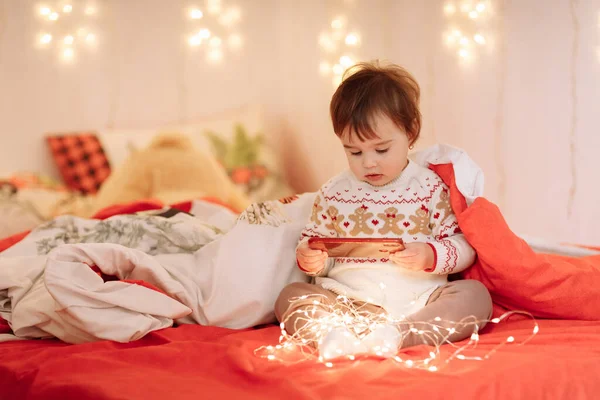 冬の装飾が施されたワーム服の愛らしい女の子は 居心地の良い寝室の装飾されたガーランド 新年の家族の休日に毛布の下のベッドで楽しく コミュニケーションし 秘密を共有しています — ストック写真