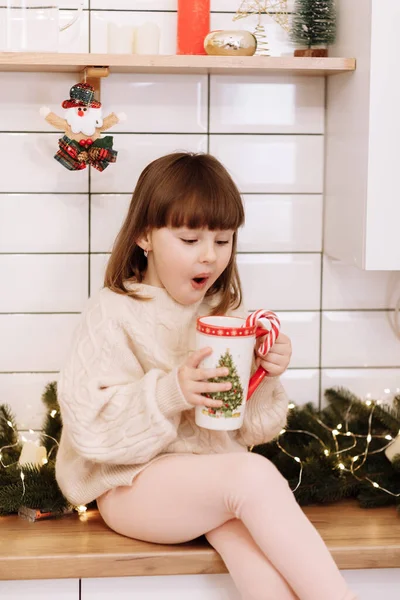 驚きの少女は 暑い飲み物とお菓子の杖でカップを開催しています休日のための装飾されたキッチンで新年を待ちます 自宅で居心地の良い暖かい冬の日 クリスマス休暇中の幸せな家族 — ストック写真