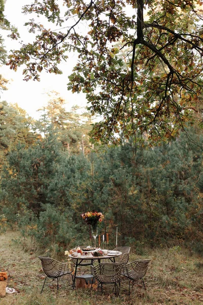 Cozy autumn picnic in the park. Festive setting table decorated wildflowers in vase, candles, food, hot drinks, maples leaves and wicker chairs outdoors. Straw baskets and pumpkins on the grass.
