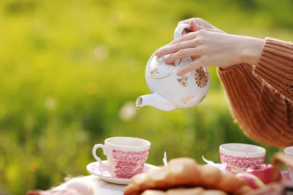 female hands pour tea into a cup from a teapot. Breakfast picnic with croissants and tea in spring blooming park on a white tablecloth on a sunny day. Outdoor, picnic, brunch, spring mood.