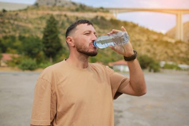 Sakallı, plastik şişeden su içen ve sıcak yaz gününde dağ manzarasının tadını çıkaran kendine güvenen genç yakışıklı bir adam. Sağlık sigortası konsepti. Sağlıklı bir yaşam tarzı. Tazeleyici içecek..