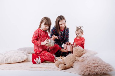 Mutlu aile kucaklaşır ve birlikte eğlenir. Kışlık pijamalı iki küçük kızı olan gülümseyen anne beyaz arka planda sıcak ekose üzerinde oturuyor. Anneler günü, bebek günü. Yeni Yıl.