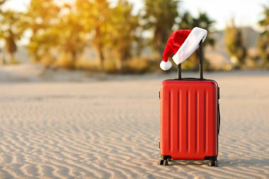 İçinde Noel Baba 'nın kırmızı şapkası olan kırmızı bavul tropikal kumsal, palmiyeler, dağlar ve deniz altyapısı olan. Noel tatili ve yeni yıl için seyahat konsepti. Mesaj için yer. İndirimli geziler.
