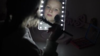 Küçük kız makyaj yapıyor ve masadaki arka farla aynaya bakıyor. Çocuk cilt okşamasına dikkat et yanaklarını fırçala evde elektrik olmadan yüzüne allık sür. Elektrik kesintisi.