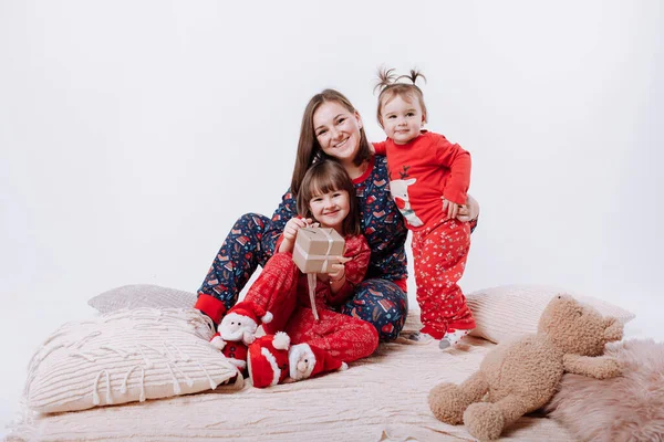 Happy family is hugging and having fun together. Smiling mom with two little daughters in winter pyjamas in xmas prints are sitting on warm plaid on white background. Mothers day, baby day. New Year.