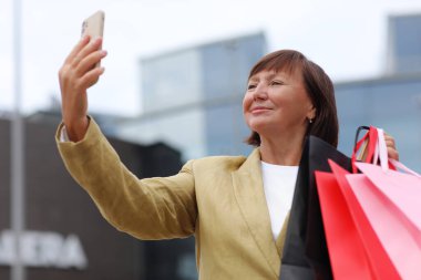 Alışveriş çantaları ve akıllı telefonları olan şık yetişkin kadın blogcu alışveriş merkezi yakınlarındaki sosyal ağlarda takipçilerle iletişim kuruyor. Tüketim, satış, zengin hayat, sanal hayat kavramı. Kara Cuma.
