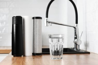 A glass of clean fresh water and set of filter cartridges on wooden table in a kitchen interior. Installation of reverse osmosis water purification system. Concept Household filtration system clipart