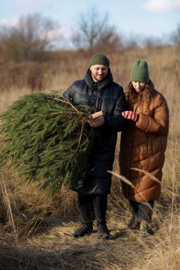 Dikey fotoğraf. Kıvırcık saçlı ve sakallı genç kadın dışarıda sarılıyor. Adam bir Noel ağacı tutuyor. Çiftler birbirlerini arıyorlar. Mutlu aile tatilleri..