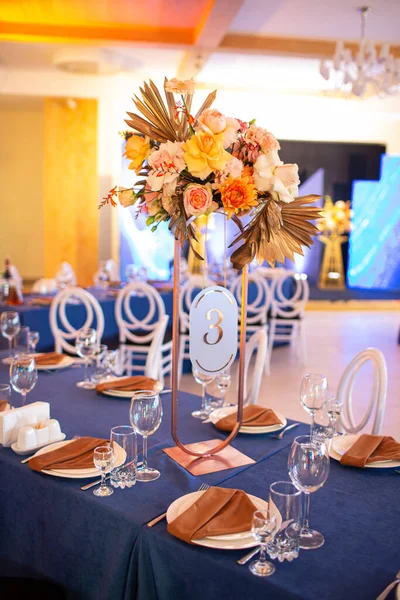 Festive table with blue cloth, napkins, plates, cutlery and glasses prepared for wedding guests in restaurant. Beautiful floral arrangement in gold metal vase with number three. Amazing servings.