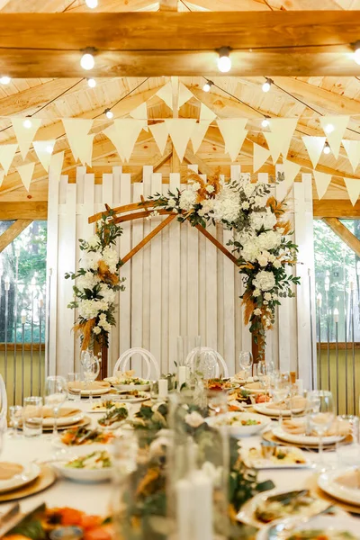 Festive setting table with meals for wedding, wooden arch, stands decorated with composition of white flowers and greenery in the rustic banquet hall