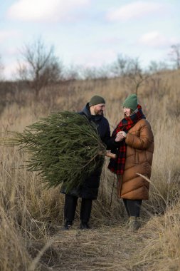 Dikey fotoğraf. Kıvırcık saçlı ve sakallı genç kadın dışarıda sarılıyor. Adam bir Noel ağacı tutuyor. Çiftler birbirlerini arıyorlar. Mutlu aile tatilleri..