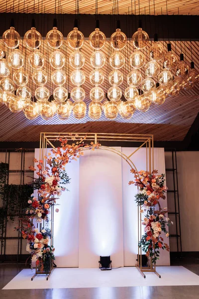 Beautiful wedding gold metal arch, wall, photo zone, area decorated with bright flowers and greens in hall for wedding ceremony. Festive decor in banquet area at party. Decorations in luxury ceremony.