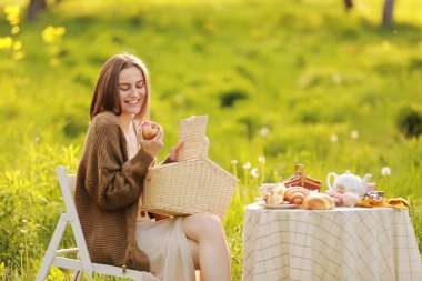 Hasır sepeti doğada yemek taşıyan genç bir kadın. Kız yazın piknik yapıyor ve parkta elma yiyor. Sağlıklı yiyecekler. İnsanlar, yaşam tarzı, rahatlama ve tatil kavramı