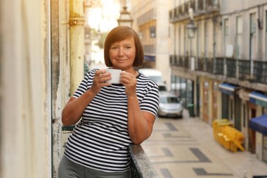 Orta yaşlı bir kadın elinde kahve ya da çay ile Avrupa şehir manzaralı bir balkonda dikiliyor. Yetişkin bir kadın tatilin tadını çıkarıyor. Yaz, hafta sonu, turizm, seyahat.