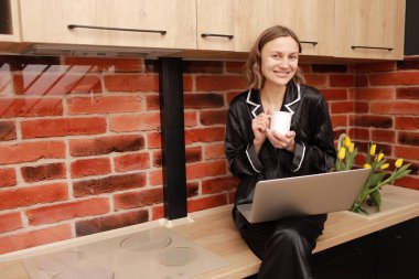 Attractive smiling young woman in pyjama drinking coffee or tea during her breakfast and working with laptop at home. Girl sits on countertop in kitchen. SMM manager enjoying working at home