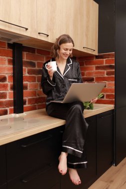 Beautiful young woman in stylish pyjama drinking coffee or tea during her breakfast and working with laptop at home. Smiling girl sits on countertop in kitchen. SMM manager enjoying working at home