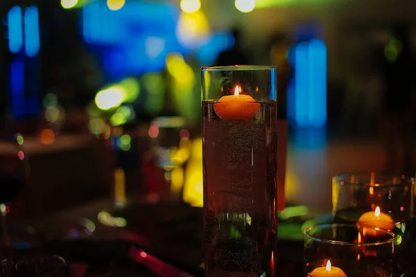Banquet table decorated with burning candles in glass vases in restaurant hall. In the background party with silhouettes of people dancing on the dance floor with disco lights glowing searchlight.