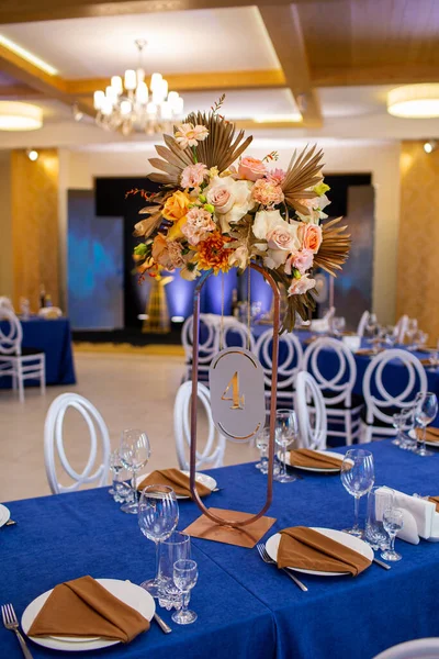 Festive table with blue cloth, napkins, plates, cutlery and glasses prepared for guests in restaurant. Beautiful floral arrangement in gold metal vase with number four. Amazing servings.