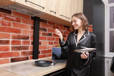 Happy smiling attractive young woman housewife preparing and cooking food and testing some meal from the frying pan on induction stove for delicious dinner at modern loft style kitchen. Advertising