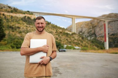 Smiling man in casual clothes standing with laptop outdoors on bridge construction background. Concept of remote work engineer or freelancer lifestyle. Cellular network broadband coverage.internet 5G.