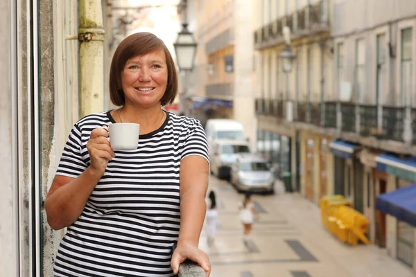Orta yaşlı bir kadın elinde kahve ya da çay ile Avrupa şehir manzaralı bir balkonda dikiliyor. Yetişkin bir kadın tatilin tadını çıkarıyor. Yaz, hafta sonu, turizm, seyahat.