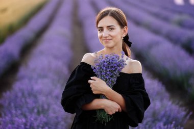 woman day. Young romantic woman with healthy natural beauty skin holding bouquet in lavender field looking at camera and smiling. Beautiful girl wearing black dress and bow on hair. Advertising natural product.