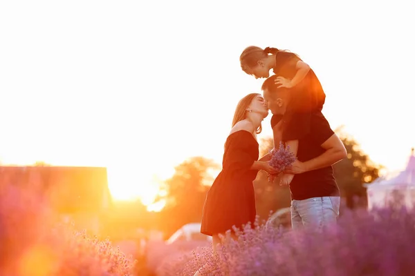 happy family day. Young mother and father carrying on shoulders daughter in lavender field on sunset. Dad, mom and child girl kissing and hugging on nature on summer. friendly family. family look.