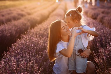 Mothers day. Young mom with little child daughter doing gesture heart with hands, hugging and kissing in a blooming lavender field. Family of two having fun, playing in meadow field on summer holiday.