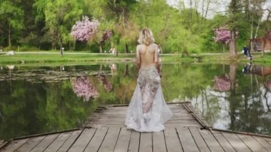 Young beautiful smiling blond woman with short hair in luxury long lace dress with crystals outdoors. Sexy happy bride dancing by the pond in blooming sakura or cherry park. International women's day.