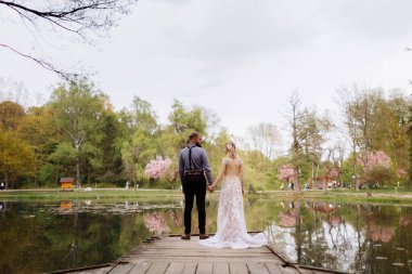 Side view of beautiful stylish newlyweds couple, bearded groom and bride in luxury long white dress are hugging by the pond in blooming pink cherry, sakura blossom park. Wedding day, valentines day.