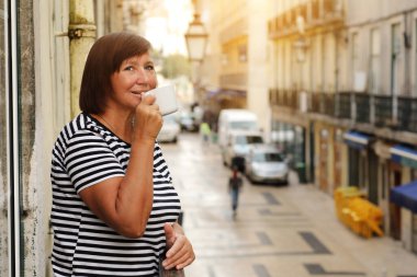 Orta yaşlı bir kadın elinde kahve ya da çay ile Avrupa şehir manzaralı bir balkonda dikiliyor. Yetişkin bir kadın tatilin tadını çıkarıyor. Yaz, hafta sonu, turizm, seyahat.