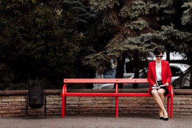 Attractive businesswoman is doing notes in paper notebook on bench in the park. Executive in red blazer and lether skirt is resolving work issues during the lunch break outdoors