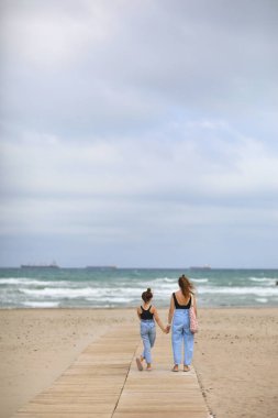 Plaj tatilindeki mutlu aile. Anne ve küçük kızın arka görüntüsü birlikte yürüyor ve sahilde el ele tutuşuyorlar. Anne ve kız çocuk yaz tatillerinde eğleniyor ve eğleniyorlar..