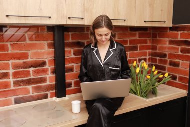 Attractive smiling young woman in pyjama drinking coffee or tea during her breakfast and working with laptop at home. Girl sits on countertop in kitchen. SMM manager enjoying working at home