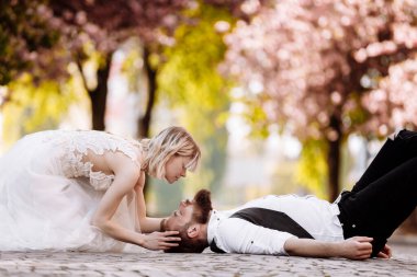 Styilish bearded man is lying in the middle of the street and woman trying to kiss him in the park with blooming cherry or sakura on background. Newlywed couple in love on wedding day, honeymoon