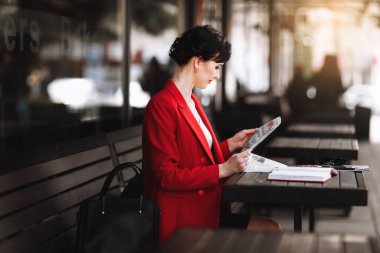 A attractive businesswoman in corporate outfit red blazer holding morning newspaper while sitting on cafe terrace. Manager is reading the latest world news during a coffee break at work.