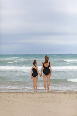 Plaj tatilindeki mutlu aile. Anne ve küçük kızın eşofmanlı halleri bir arada ve plajda el ele tutuşuyorlar. Anne ve kız çocuk yaz tatillerinde eğleniyor ve eğleniyorlar..