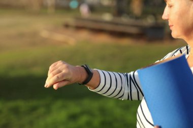 Dünya Sağlık Günü. Olgun bir kadın yoga minderini tutuyor ve her sabah doğa rutinini kontrol etmek için akıllı saatlere bakıyor. Yaşlılar için sağlıklı yaşam tarzı, canlılık, sağlık hizmeti kavramı.