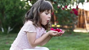 Adorable little child girl 5-6 years old is blowing red roses petals in hands outdoors on summer day. Kid having fun at home backyard with green grass and big roses bush on background. Children day.